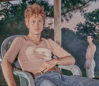 A white man wearing a beige top and baggy jeans louges in a plastic chair.