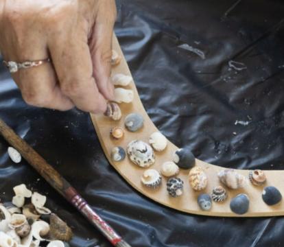 Two hands are placing small shells onto a light brown boomerang.