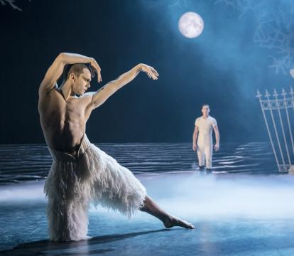 A topless man in a white feathery costume dancing under a set with a full moon on a blue lit stage.