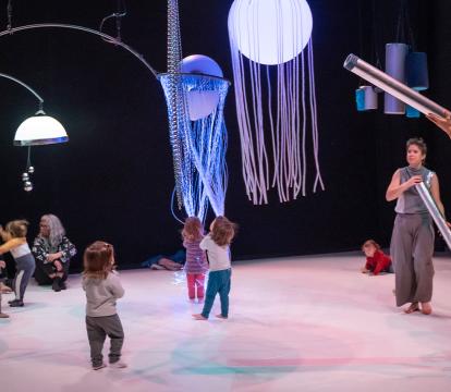 A group of young children plays under jellyfish-like lights on a stage, with adults engaging them using tubular instruments.