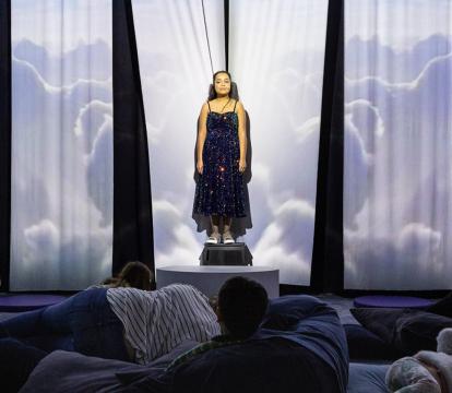 A young white woman with dark brown hair stands in front of a fabric background, where projections of clouds are being cast. An audience of meditation children and adults lie on pillows in front of her.
