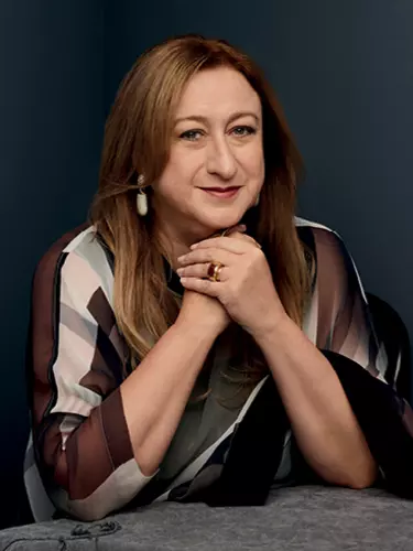 A white woman in her 60s with long brown hair holds her hands under her chin looking at the camera with a small, warm smile.