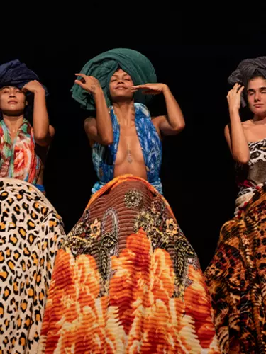 Three caramel-skinned female dancers are mid-dance move. They wear towel-looking headpieces and have colourful patterned skirts.