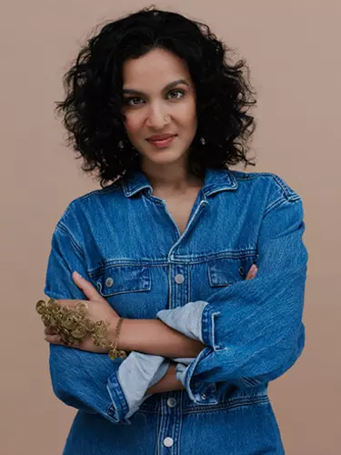 An Indian woman with short, curly black hair looking directly at the camera wears a denim shirt with gold hand jewellery and a nose ring.