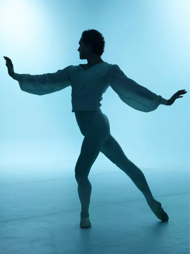 A male ballet dancer in silhouette wears a flowy shirt and his arms are outstretched, one in front of him and one behind.