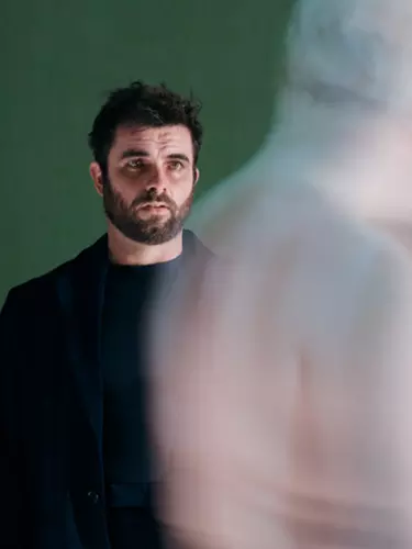 A white man with black hair and dark beard wears all black. He's in front of a green background and is looking at a shadow.