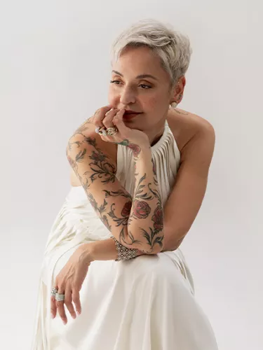 A woman adorned with tattoos with short, bleached blonde hair is comfortably positioned on a chair, resting her chin in her hand.