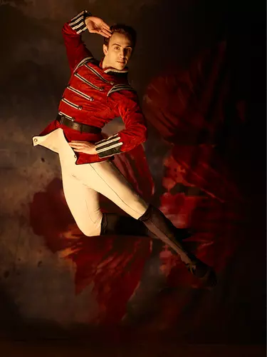 A white male ballerina with brown hair wears a toy soldier's costume with red jacket, light beige pants and dark brown boots. He is mid-jump and is saluting.