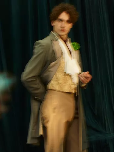 A blurry image of a man standing straight. He has brown curly hair and wears a gold vest, long green jacket and light brown pants.