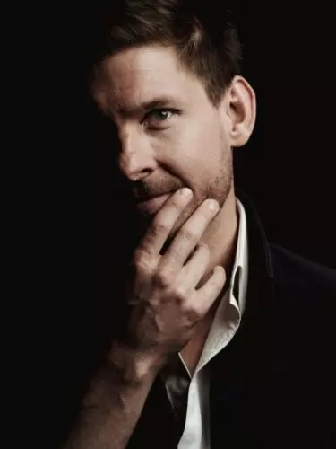 A close up of a white 30-40 year old man wearing a suit has his hand on his chin. Half his face is in darkness.