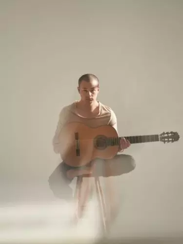 A young Asian man with shaved hair wears a beige top and holds an acoustic guitar.