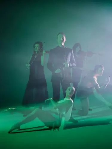 A white, 50-year old man holds a violin immersed in green light. Around him is two female violinists, a man dancer and a female ballerina.