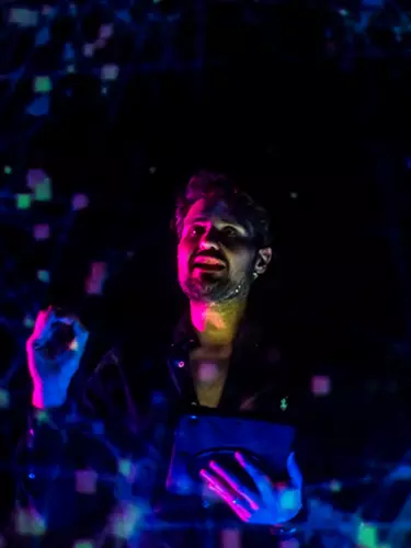 A white man with short brown hair is conducting. There are pink and blue lights on his face.