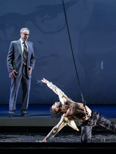 A white man with grey hair in a suit stands looking at a male dancer knelt on the floor. The dancer wears a harness and is dancing in water.