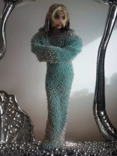 A person dressed in a long, blue beaded gown with a unique headpiece, standing within an abstract metallic frame.