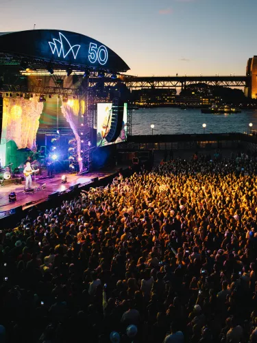 A large crowd stands in front of an outdoor stage at sunset with the Sydney Harbour Bridge in the background.