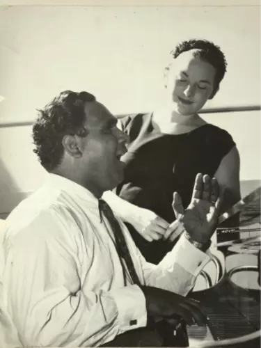 A man sitting at a desk talking to a woman who is standing nearby behind him.