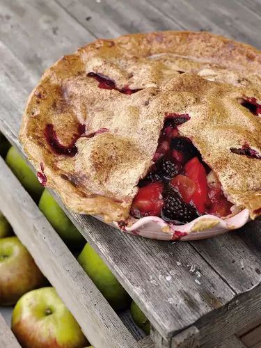Blackberry and apple pie in a white dish with a slice cut out of the pastry, sitting on a wooden pallet.