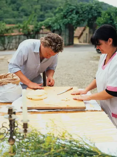 Jamie Oliver and another person standing around a table, rolling pasta out on a wooden chopping board in an outdoor setting.