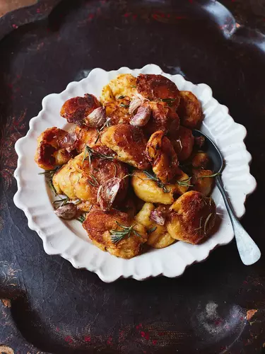 A pile of roasted potatoes on a white plate on a dark tabletop, with a silver spoon resting on the side of the plate.