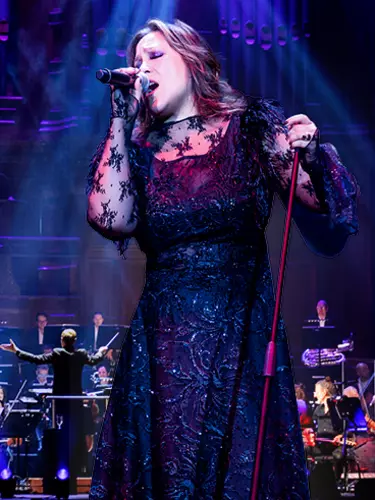 A Filipino-Australian woman with brown layered, shoulder-length hair holds a microphone in her right hand and the microphone stand in her left hand. She's singing on stage in front of an orchestra.