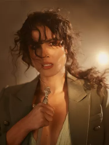 A white woman with a messy, curly ponytail and wearing a beige blazer looks worried at the camera while holding the handle of a sword.