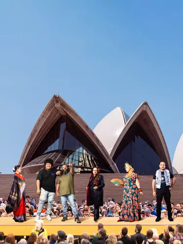 Three men and three women from different backgrounds stand on a yellow platform in front of the Sydney Opera House sails in front of an audience.