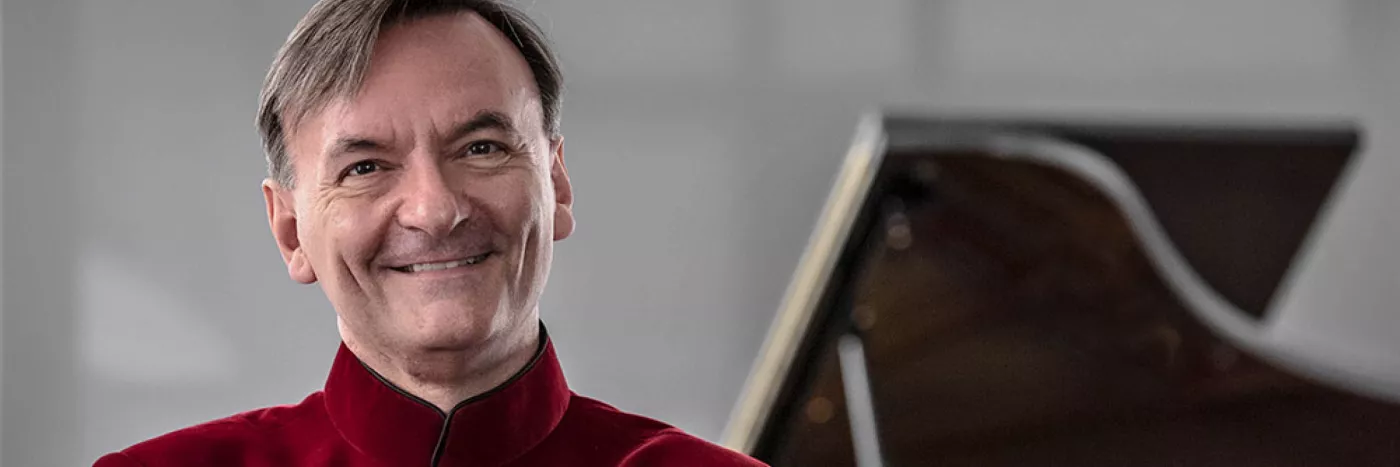 Stephen Hough wears a red shirt standing in front of a grand piano
