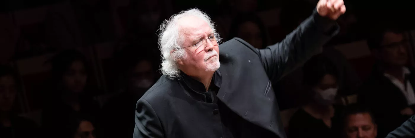 Donald Runnicles stands with his left hand raised as he conducts an orchestra