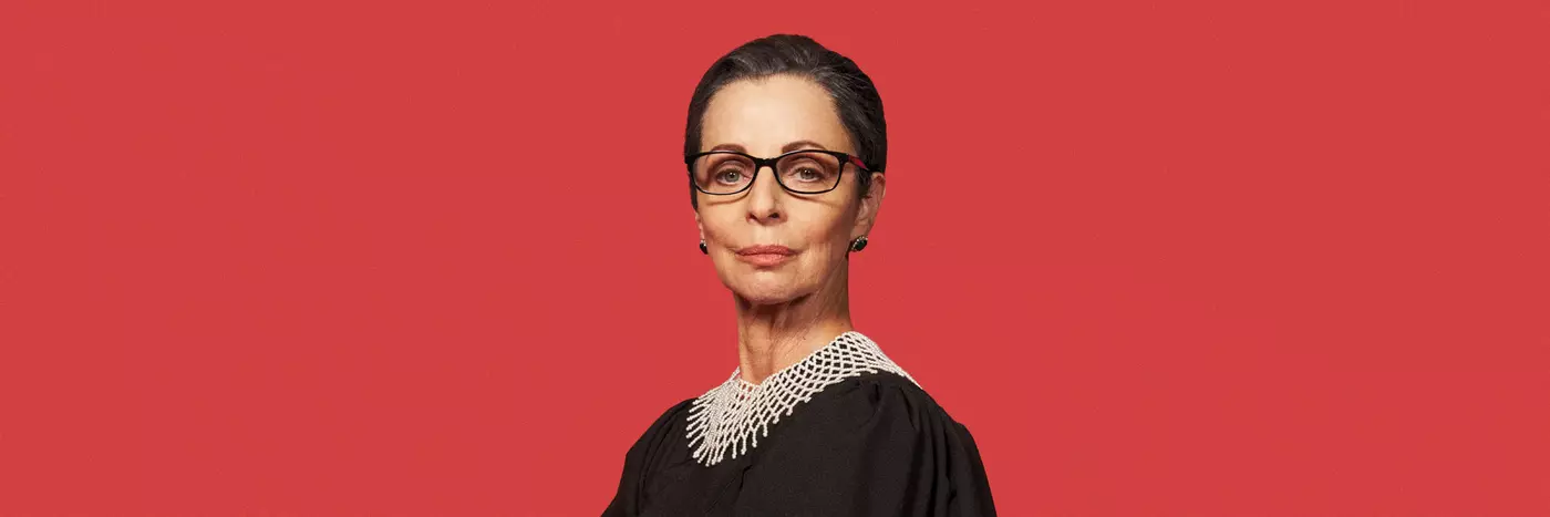 A white woman in her 60s has her hair sleekly pulled back and pursed lips wears black glasses and a lawyer's black robe with white crocheted neck piece. She stands in front of an all-red background.