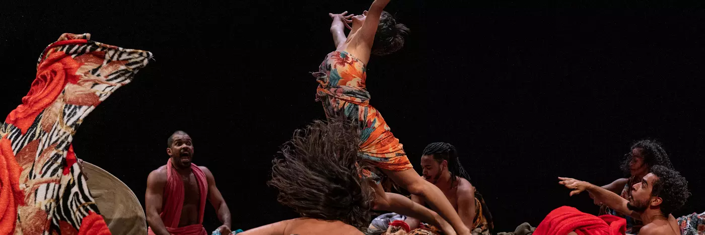 A group of Brazilian dancers, men and women, wear clothes with red and animal print patterns. They are on stage and a female dancer is mid-air as if just thrown by the male dancers.