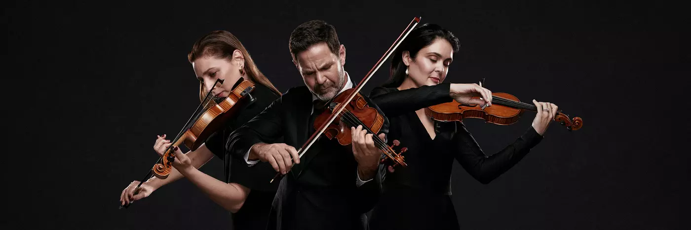 Three violinists (one man and two women) play violin with intensity, in front of a black backdrop.