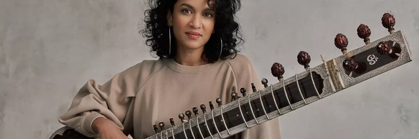 An Indian woman with short, curly black hair wears big silver hoop earrings and a beige long sleeve top. She has a sitar placed across her knee - this is a guitar-like instrument with a short round body and a very long neck and many tuning knobs.