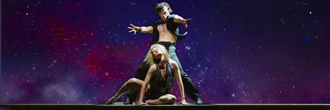 A male dancer wearing black and an open button shirt stands behind a female dancer sitting on the floor.