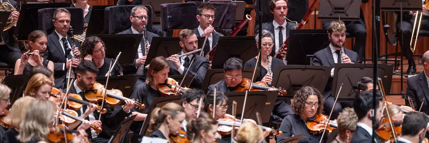 An orchestra performing. We see men and women playing a variety of instruments including violins, oboes, trumpets and flutes.