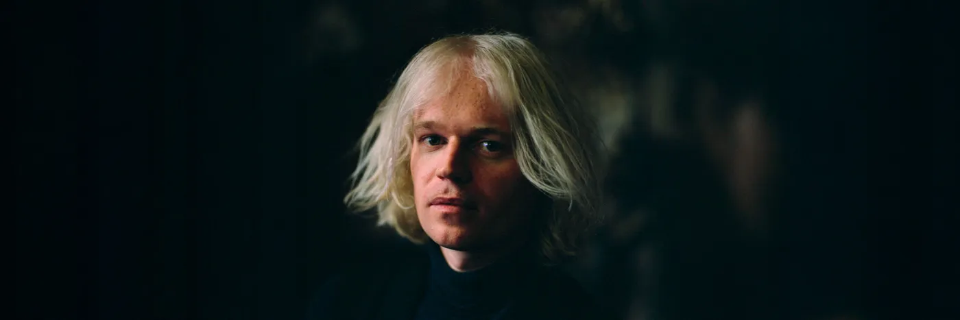 A white man in early 40s with jaw-length white hair looks at the camera.