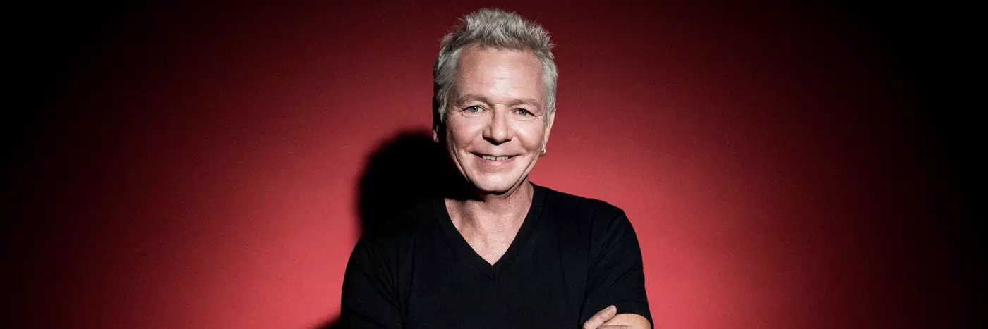 A white man in his 60s with grey hair wears a black t-shirt and has his arms crossed, standing in front of a red background.