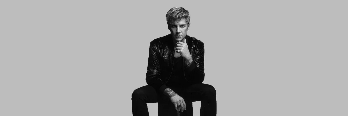 A black and white image of a white man with blonde, spikey hair sitting on a stool wearing a leather jacket