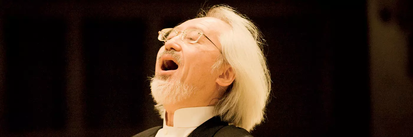 A man with white hairs and in black suit singing on the stage.