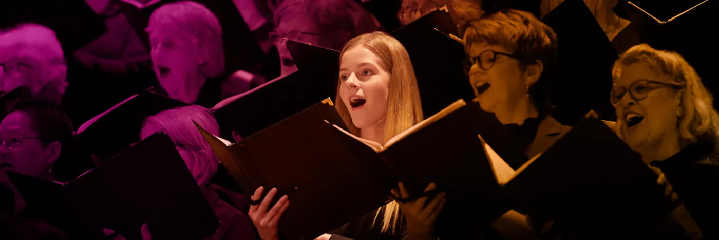 A blonde girl with an open song book sings amongst a choir