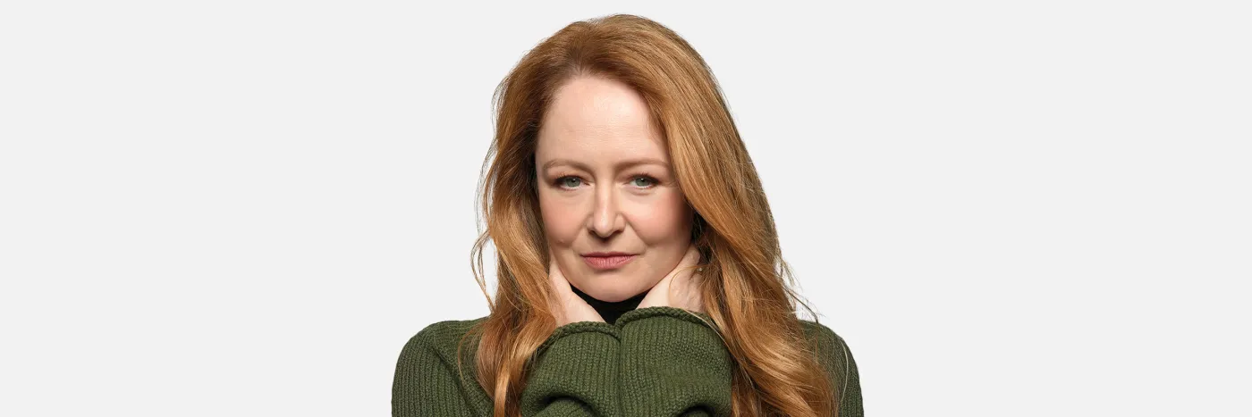 A woman with long red hair wearing a green sweater, holding her hands up under her face against a white background.