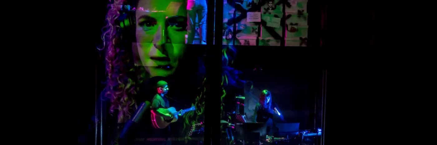 A man wearing a beanie plays the guitar while a woman next to him plays a keyboard, They are surrounded by a projection of a woman's face, and the lighting is green, purple and blue.