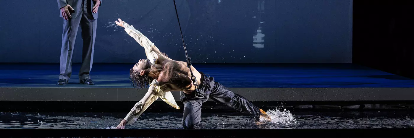 A white man with grey hair in a suit stands looking at a male dancer knelt on the floor. The dancer wears a harness and is dancing in water.