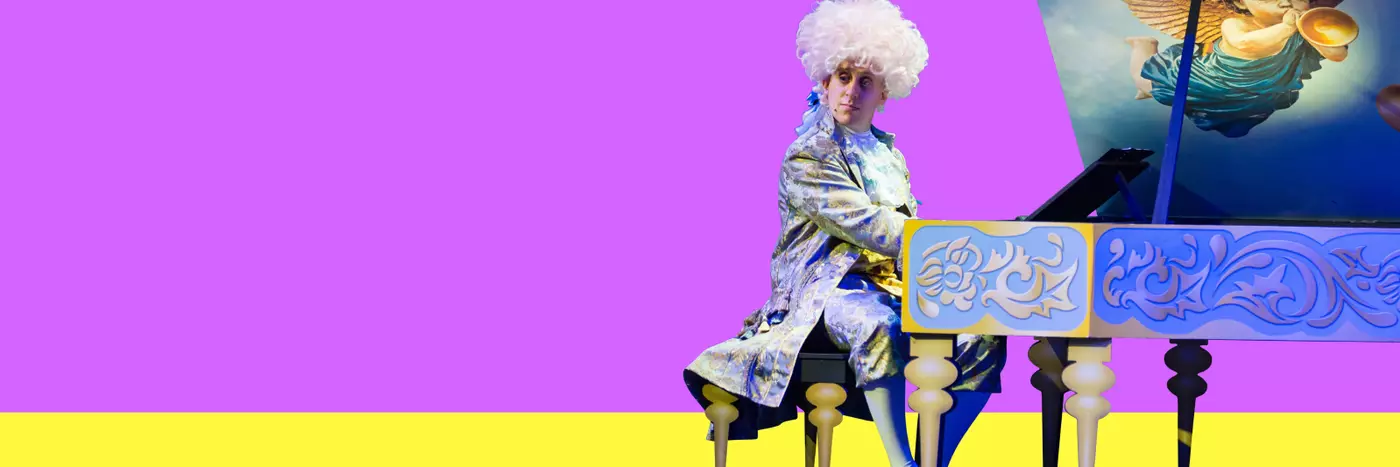 A man wearing a Mozart costume and a big white wig sitting at a grand piano