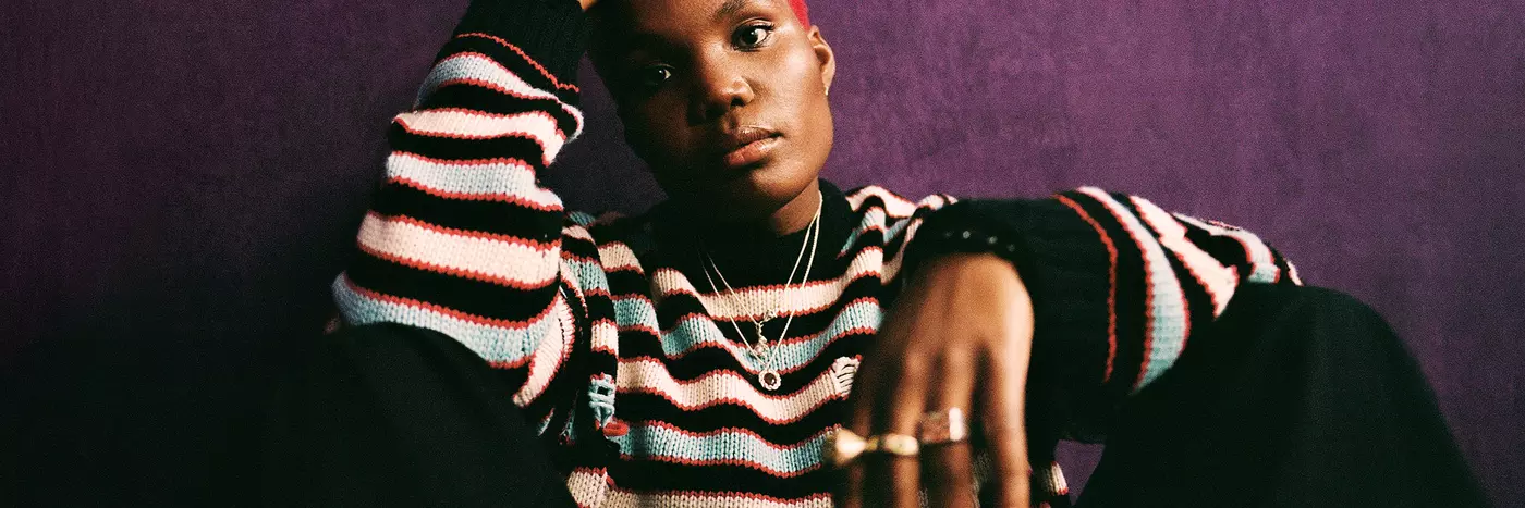 Arlo Parks wearing a striped knitted sweater, sitting on the ground against a dark purple wall.