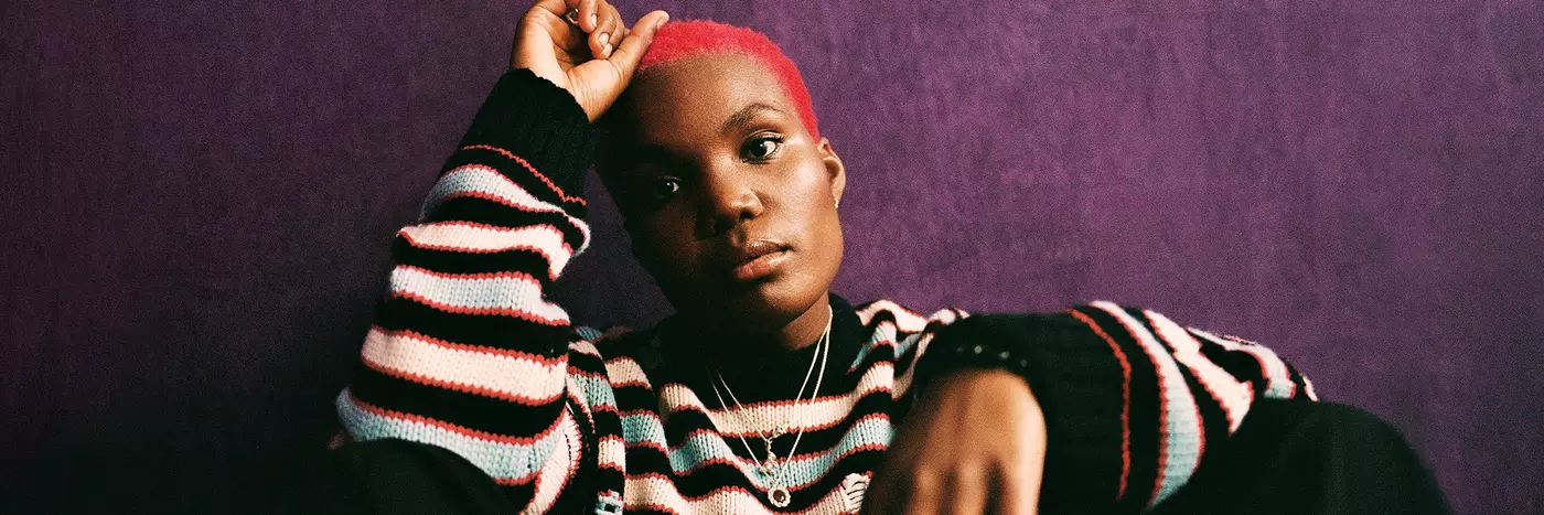 Arlo Parks wearing a striped knitted sweater, sitting on the ground against a dark purple wall.