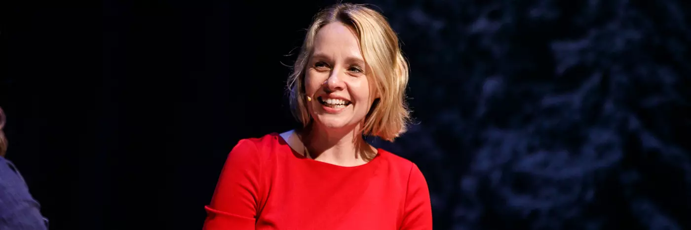 A person in a red t-shirt sitting on stage and smiling as if in conversation.