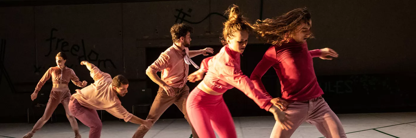 Five dancers wear pink and stand in a diagonal line caught in movement.