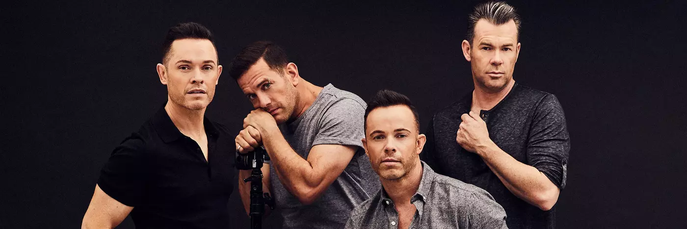 Four white men wearing various shades of black, white and grey are posed in front of a black background. Three men are standing and one is seated on a stool.