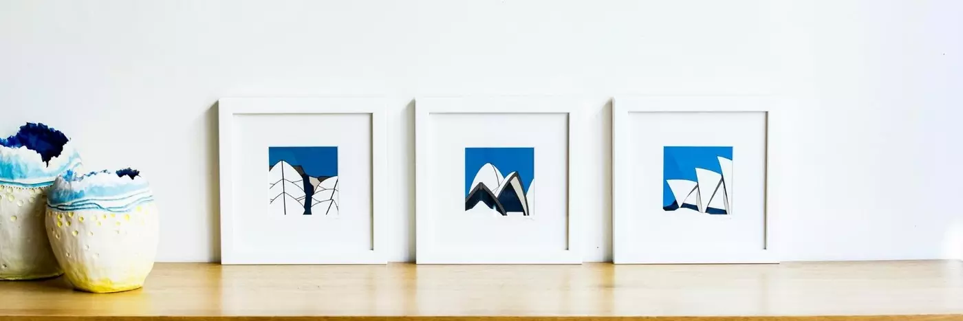 Three framed blue pictures resting on a wooden cabinet against a white wall.